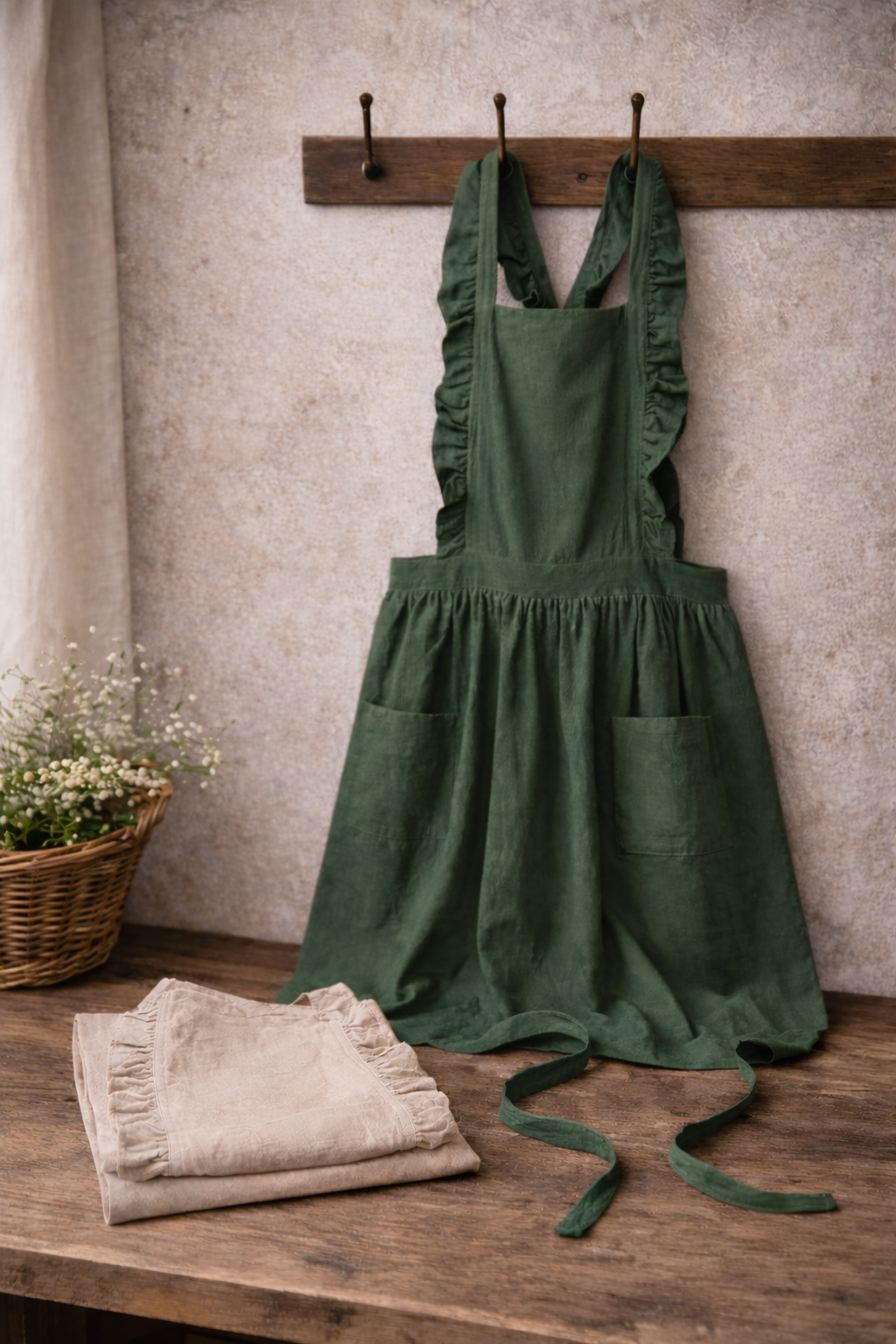 Grönt linneförkläde hänger över krokar med naturliga veck, beige förkläde vikt på träbord i lantlig miljö